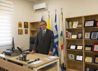 ΑΝΑΚΟΙΝΩΣΗ: 12-09-2024 Ανδρας σε γραφείο με ελληνικές σημαίες και βραβεία, προωθεί τη δημοκρατία και την πολιτική συμμετοχή.