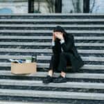 ΑΝΕΡΓΙΑ-ΡΕΚΟΡ ΣΤΙΣ ΓΥΝΑΙΚΕΣ ΚΑΙ ΤΟΥΣ ΝΕΟΥΣ – Η ΚΥΚΛΙΚΗ ΔΗΜΟΚΡΑΤΙΑ ΑΠΑΙΤΕΙ ΑΜΕΣΕΣ ΛΥΣΕΙΣ! Άνεργη γυναίκα καθισμένη σε σκαλιά με κουτί αποκατοίκηση, συμβολισμός οικονομικής δυσκολίας.