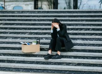 ΑΝΕΡΓΙΑ-ΡΕΚΟΡ ΣΤΙΣ ΓΥΝΑΙΚΕΣ ΚΑΙ ΤΟΥΣ ΝΕΟΥΣ – Η ΚΥΚΛΙΚΗ ΔΗΜΟΚΡΑΤΙΑ ΑΠΑΙΤΕΙ ΑΜΕΣΕΣ ΛΥΣΕΙΣ! Άνεργη γυναίκα καθισμένη σε σκαλιά με κουτί αποκατοίκηση, συμβολισμός οικονομικής δυσκολίας.