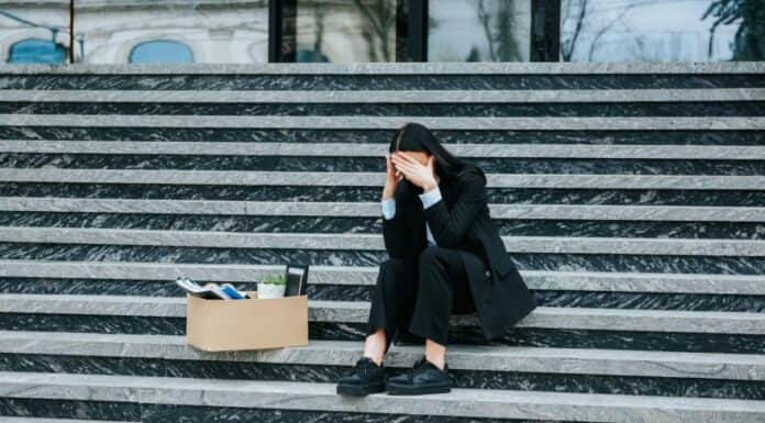 ΑΝΕΡΓΙΑ-ΡΕΚΟΡ ΣΤΙΣ ΓΥΝΑΙΚΕΣ ΚΑΙ ΤΟΥΣ ΝΕΟΥΣ – Η ΚΥΚΛΙΚΗ ΔΗΜΟΚΡΑΤΙΑ ΑΠΑΙΤΕΙ ΑΜΕΣΕΣ ΛΥΣΕΙΣ! Άνεργη γυναίκα καθισμένη σε σκαλιά με κουτί αποκατοίκηση, συμβολισμός οικονομικής δυσκολίας.