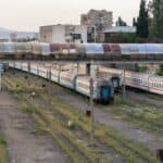 Η ΚΥΒΕΡΝΗΣΗ ΠΟΥΛΑΕΙ “ΤΡΕΝΑ” ΤΟΥ 2027, ΕΝΩ Η ΕΛΛΑΔΑ ΤΟΥ 2025 ΒΟΥΛΙΑΖΕΙ ΣΤΗΝ ΑΝΑΣΦΑΛΕΙΑ ΚΑΙ ΤΟΝ ΑΥΤΑΡΧΙΣΜΟ Οχήματα τρένων και σήραγγες σε εγκατάλειψη, εικόνα αστικού περιβάλλοντος με παλιές σιδηροδρομικές γραμμές και κτίρια.