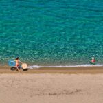 Στο σφυρί οι παραλίες μας: Η Κυβέρνηση μετατρέπει τον εθνικό μας πλούτο σε ψηφιακό εμπόρευμα! Παιδιά απολαμβάνουν το καλοκαίρι στην παραλία με σωσίβια και παιχνίδια.