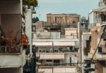 ΜΕΤΑΝΑΣΤΕΣ ΣΤΗΝ ΙΔΙΑ ΜΑΣ ΤΗ ΧΩΡΑ: 3 ΣΤΟΥΣ 10 ΕΛΛΗΝΕΣ ΕΤΟΙΜΑΖΟΥΝ ΒΑΛΙΤΣΕΣ ΤΟ 2026 Ανθρώπινη πόλη με πολυκατοικίες και αρχαίο μνημείο στο βάθος.