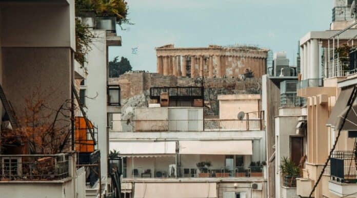 ΜΕΤΑΝΑΣΤΕΣ ΣΤΗΝ ΙΔΙΑ ΜΑΣ ΤΗ ΧΩΡΑ: 3 ΣΤΟΥΣ 10 ΕΛΛΗΝΕΣ ΕΤΟΙΜΑΖΟΥΝ ΒΑΛΙΤΣΕΣ ΤΟ 2026 Ανθρώπινη πόλη με πολυκατοικίες και αρχαίο μνημείο στο βάθος.